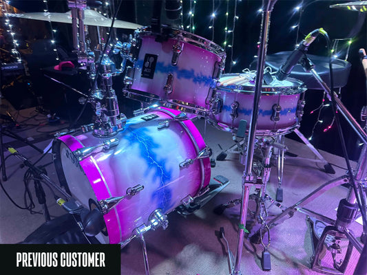 A custom drum kit with a pink and blue lightning-bolt gradient wrap, shown on a stage with microphones and string lights.